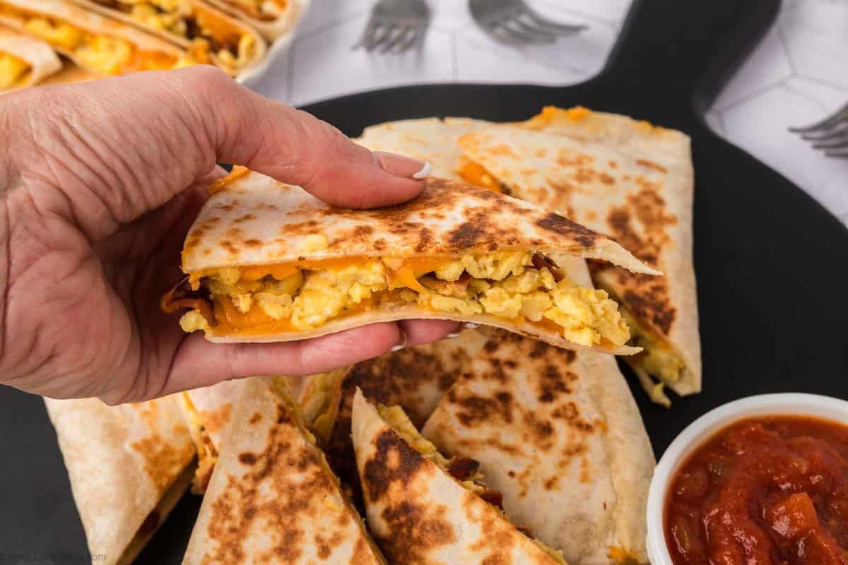 A hand holding a Breakfast Quesadilla filled with scrambled eggs and cheese, with more quesadilla slices and a small bowl of salsa on a plate in the background.