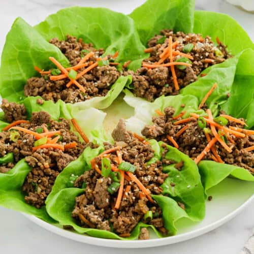 A plate of Ground Beef Lettuce Wraps filled with seasoned meat, shredded carrots, chopped green onions, and sprinkled with sesame seeds, arranged in a circular pattern.