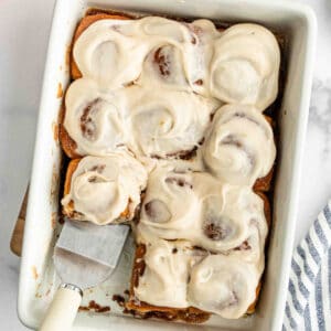 A baking dish filled with Hawaiian Roll Cinnamon Rolls topped with creamy white icing. One cinnamon roll has been cut and is being lifted with a spatula. A striped cloth is visible nearby.