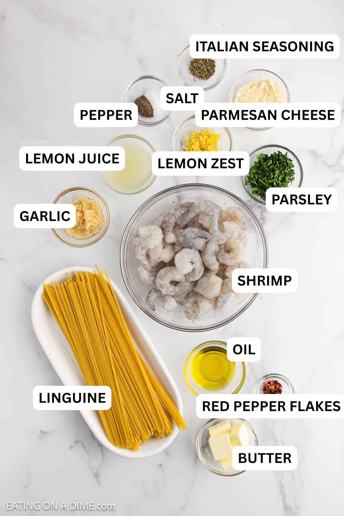 Top-down view of raw shrimp in a bowl, uncooked spaghetti, and small bowls of herbs, spices, garlic, lemon, butter, oil, Parmesan cheese, and red pepper flakes.