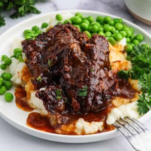 A plate of slow cooker beef ribs with rich brown sauce served over mashed potatoes, garnished with fresh parsley and accompanied by green peas. Bowls of mashed potatoes and peas are in the background.