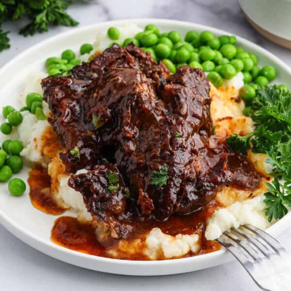 A plate of slow cooker beef ribs with rich brown sauce served over mashed potatoes, garnished with fresh parsley and accompanied by green peas. Bowls of mashed potatoes and peas are in the background.