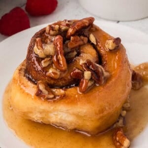 A sticky bun topped with chopped pecans and drizzled with caramel sauce sits on a white plate, with a few raspberries in the background.