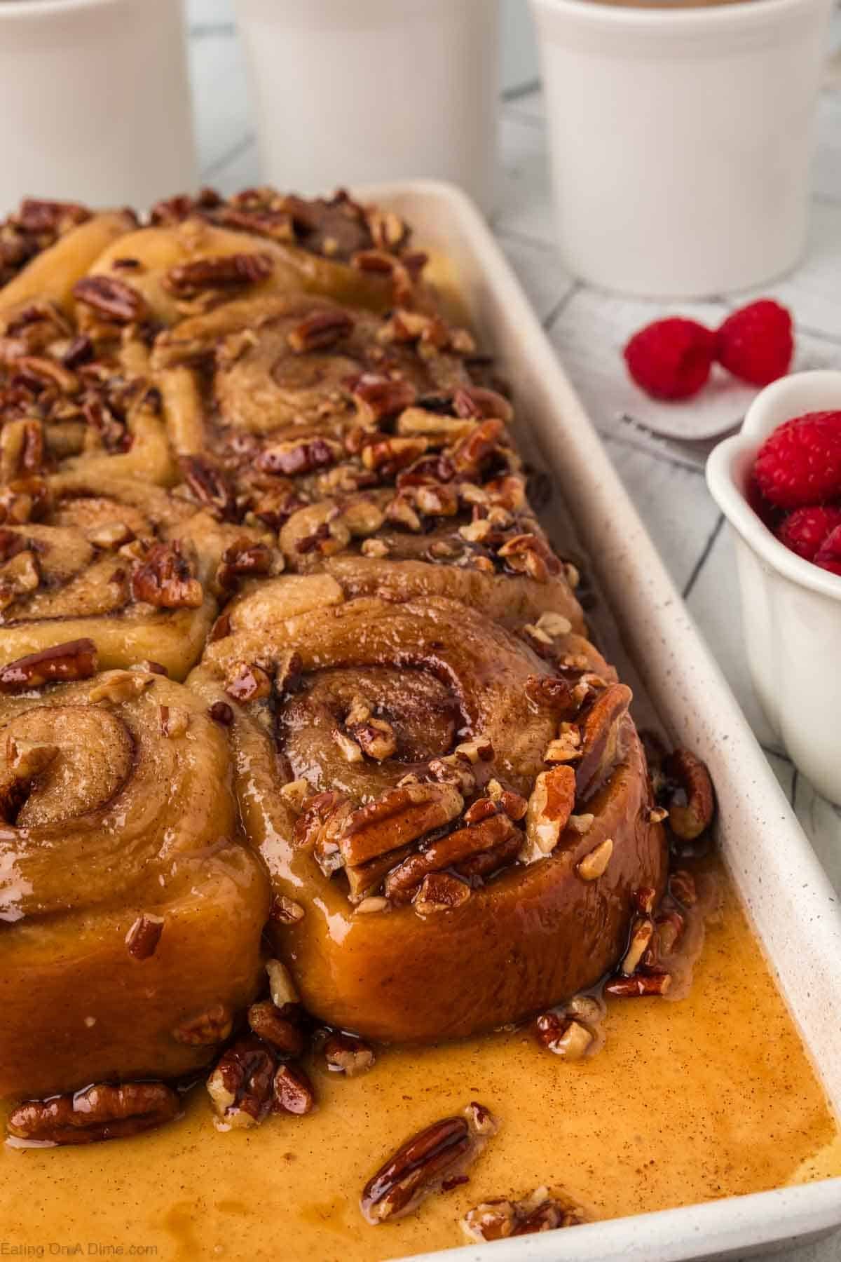 A tray of sticky pecan cinnamon rolls covered in caramel sauce, with chopped pecans scattered on top. Next to the tray is a small bowl of fresh raspberries and some white cups in the background.