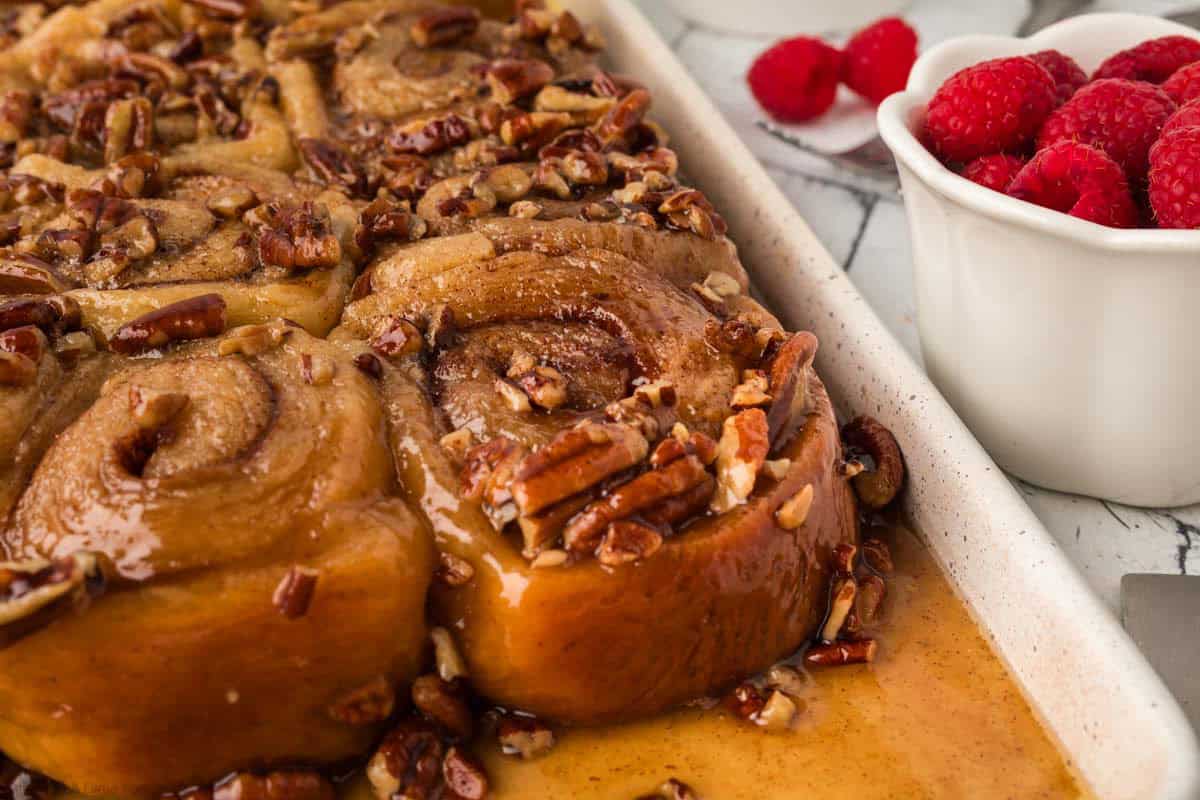 Freshly baked sticky buns topped with chopped pecans and caramel glaze sit in a baking dish next to a small white bowl filled with fresh raspberries on a marble countertop.