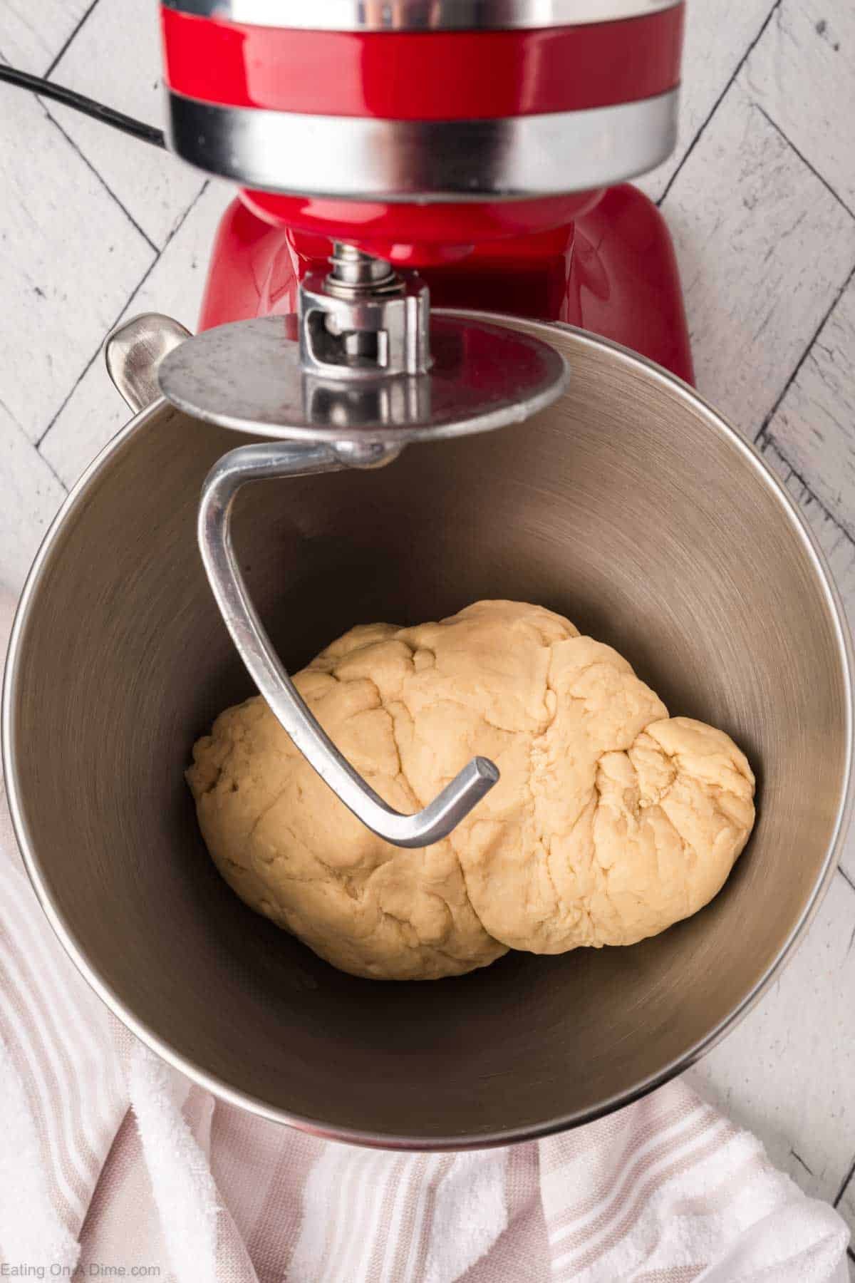 A red stand mixer with a dough hook attachment is mixing a ball of dough in a stainless steel bowl. A white and pink striped towel is partially visible underneath the mixer on a white wooden surface.