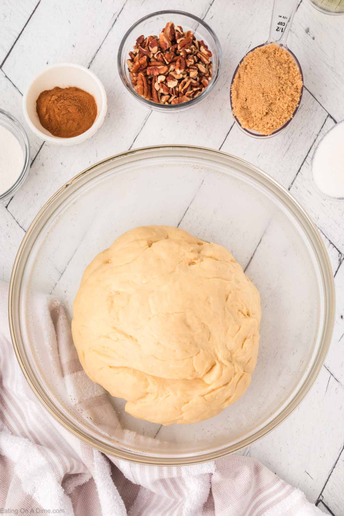 A glass bowl with a ball of dough sits on a white wooden surface, surrounded by small bowls of chopped pecans, brown sugar, cinnamon, and other baking ingredients. A striped towel is partially visible in the lower corner.