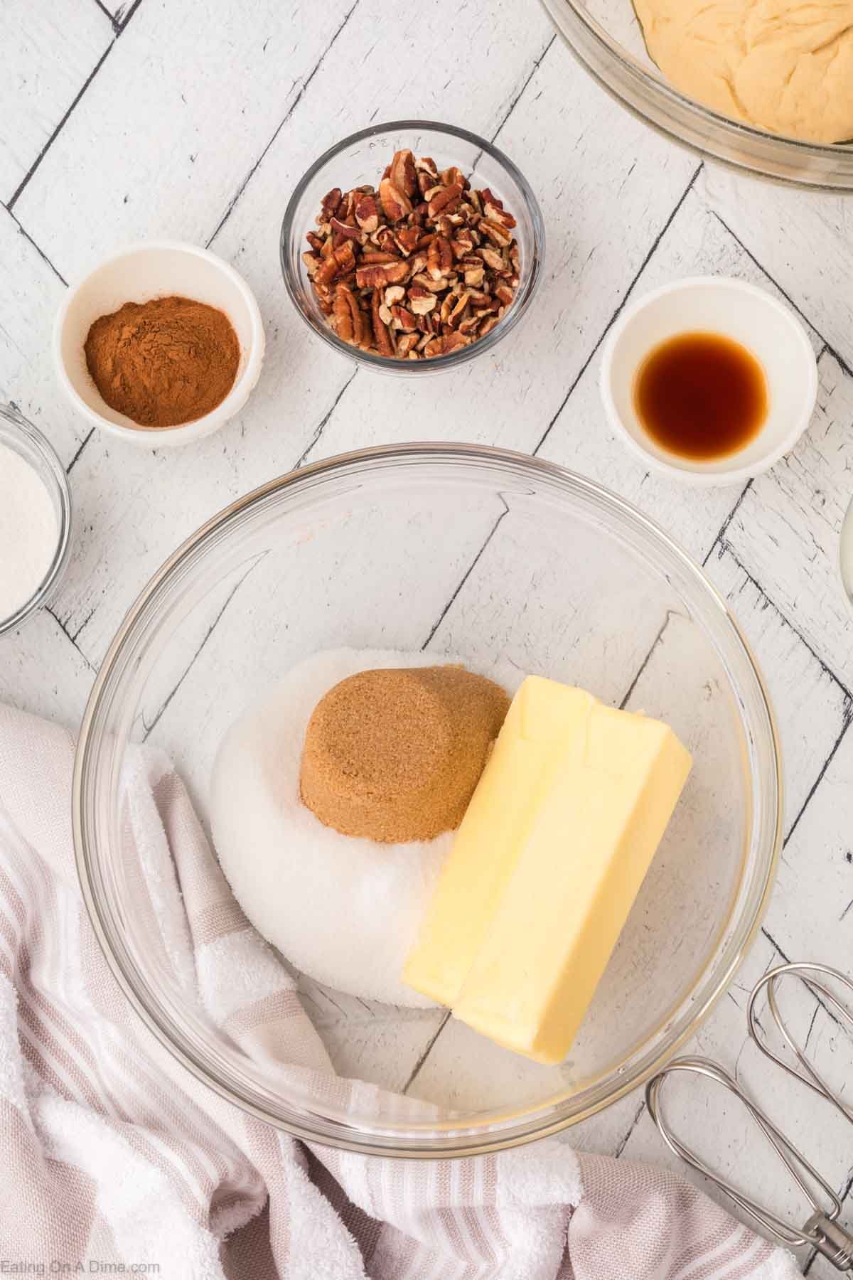 A glass bowl with butter, brown sugar, and white sugar sits on a white wooden surface. Surrounding it are small bowls of cinnamon, chopped pecans, vanilla, and a large bowl of dough. A mixer and towel are nearby.