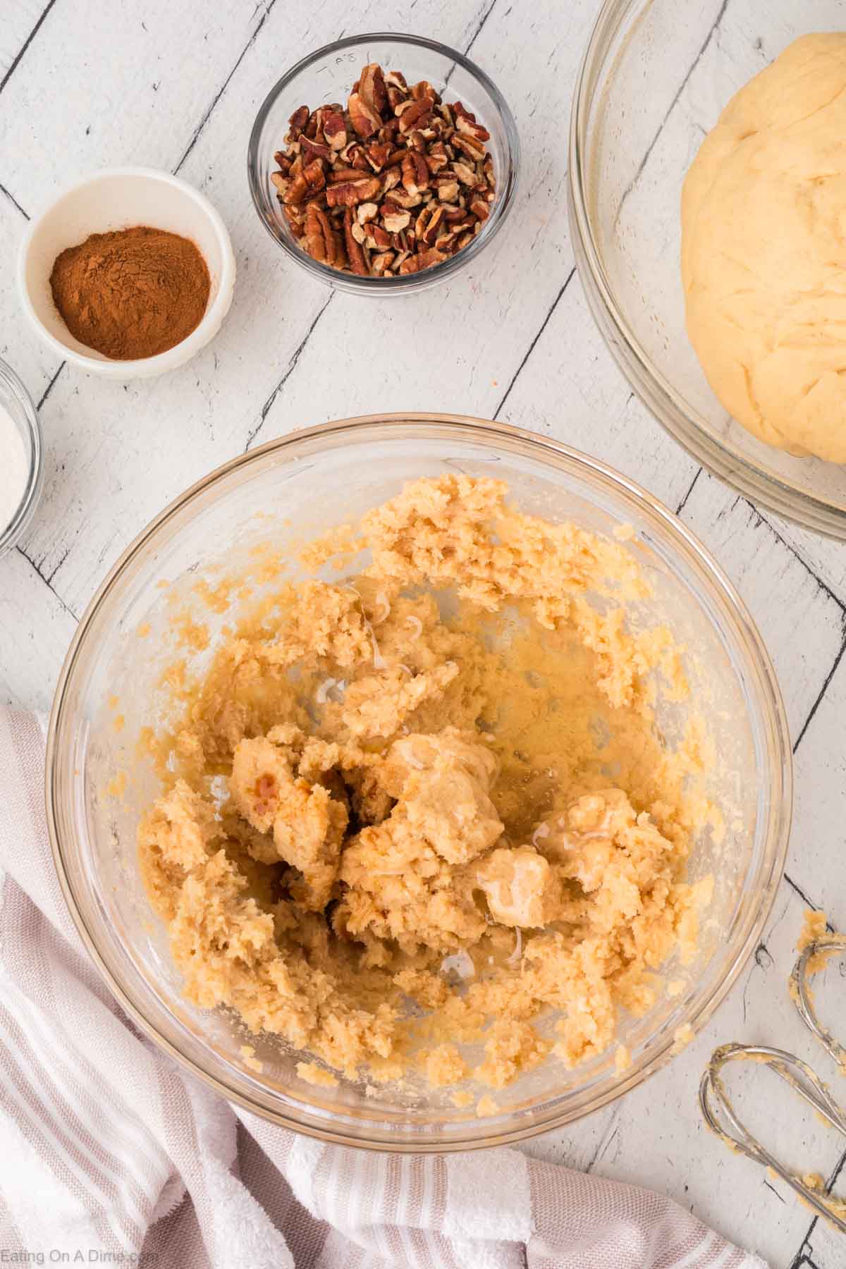 A glass bowl with wet cookie dough sits on a light wooden surface surrounded by a bowl of chopped pecans, a bowl of cinnamon, and a large bowl of dough. A hand mixer and a towel are nearby.