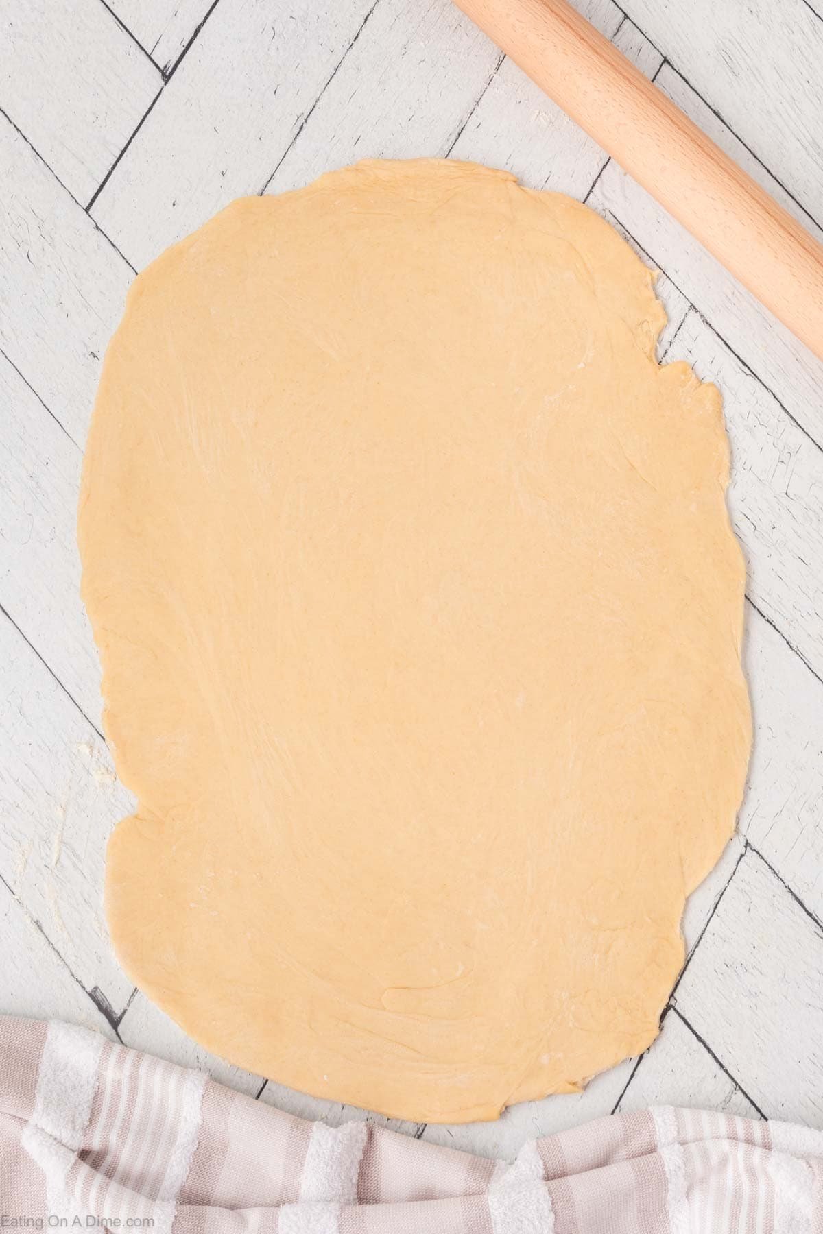 A sheet of rolled out dough on a white wooden surface, with a rolling pin above and a striped kitchen towel below.