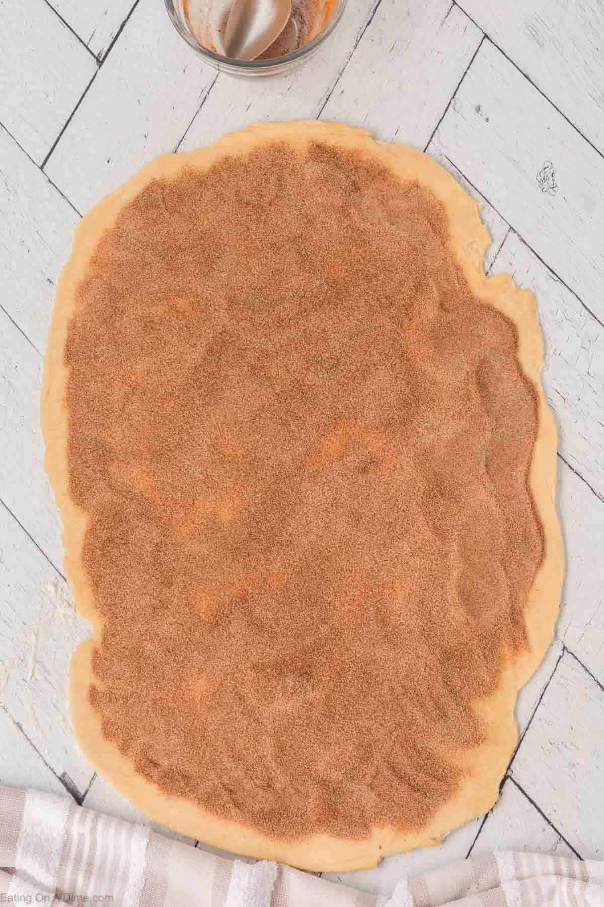 Rolled out dough on a white wooden surface, evenly covered with a layer of cinnamon sugar. A small bowl with a spatula and a striped cloth are nearby.