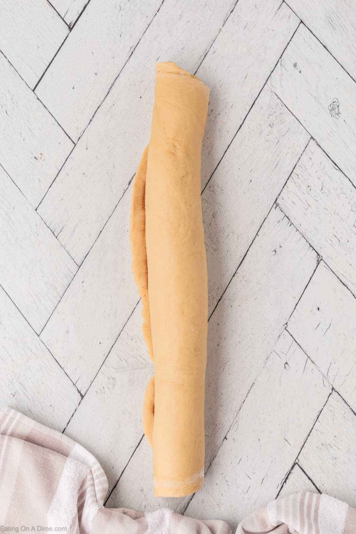 A rolled log of dough on a light-colored, herringbone-patterned surface, with a beige and white striped towel partially visible in the corner.