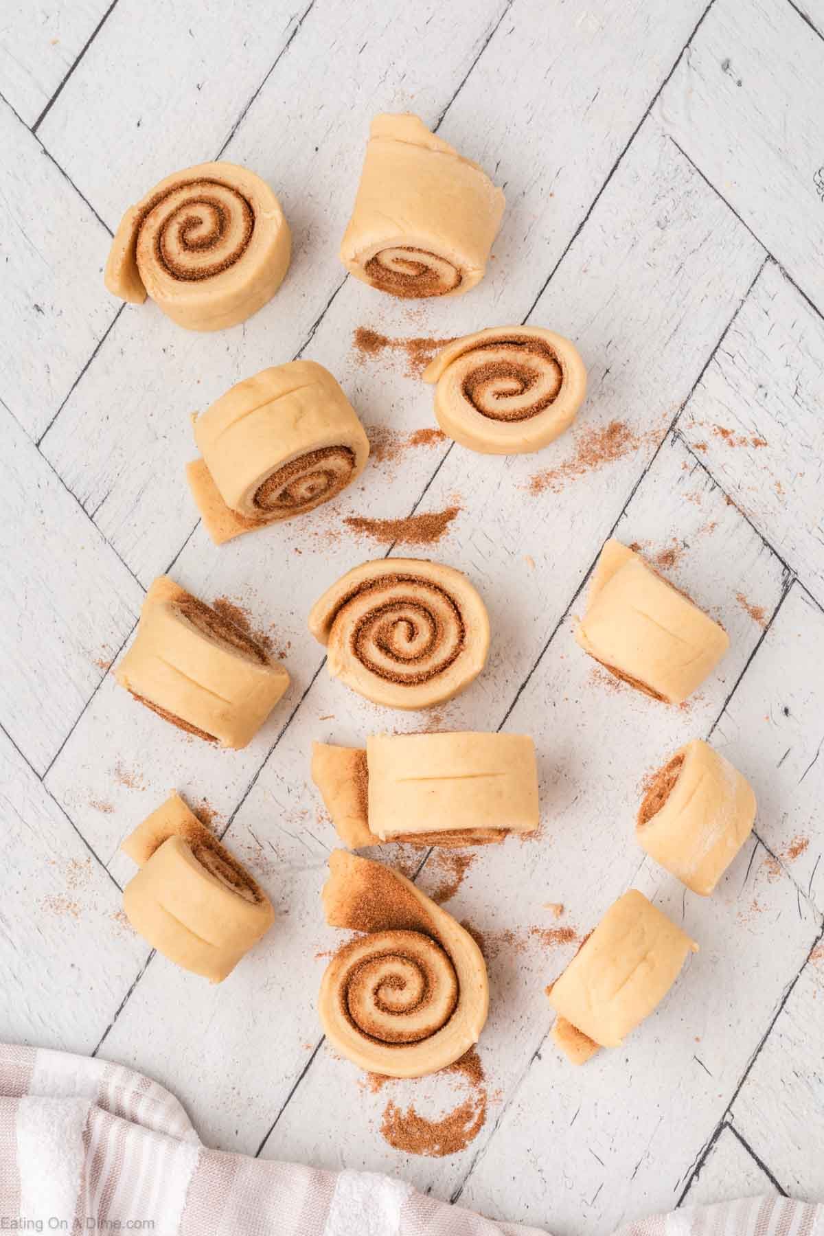 Unbaked cinnamon roll dough pieces scattered on a light, textured surface, with visible cinnamon filling and some loose cinnamon sprinkled around. A towel is partially visible at the bottom left corner.