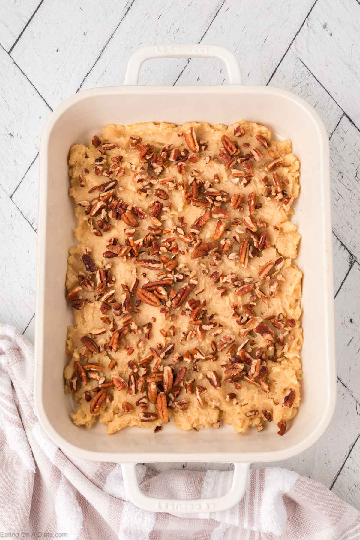 A white baking dish filled with an unbaked casserole topped with chopped pecans sits on a white wooden surface, next to a white and pink striped cloth.