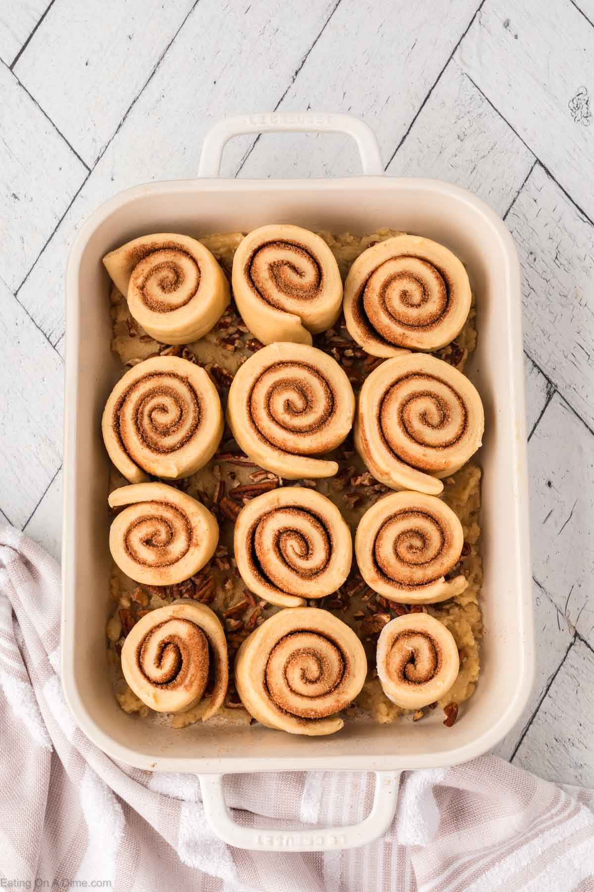 A baking dish filled with raw cinnamon rolls arranged in neat rows on top of a layer of chopped pecans, ready to be baked. A white towel is draped nearby on a light herringbone-patterned surface.
