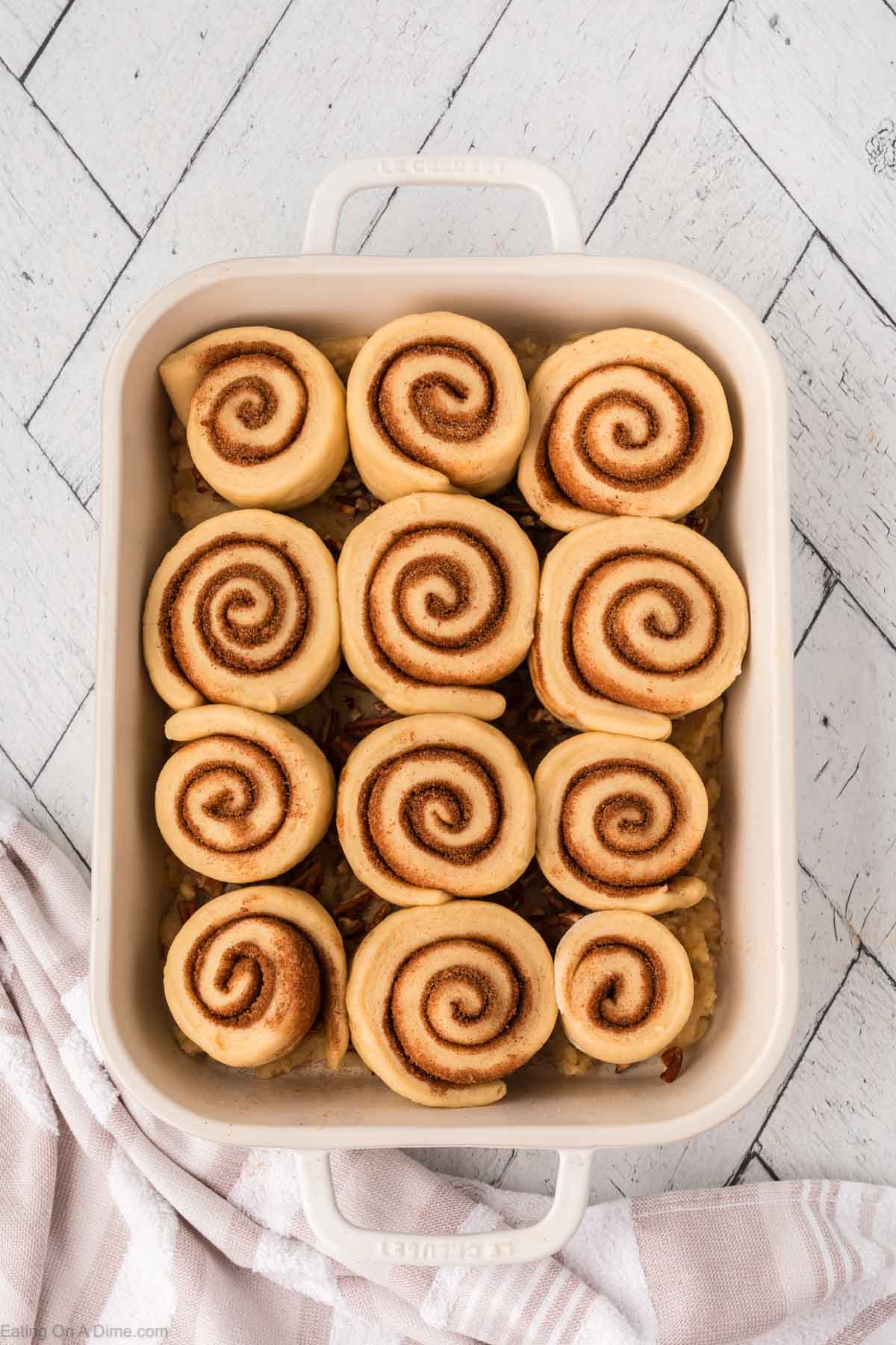 A white baking dish filled with 12 unbaked cinnamon rolls arranged in neat rows, sitting on a white wooden surface with a striped towel nearby.