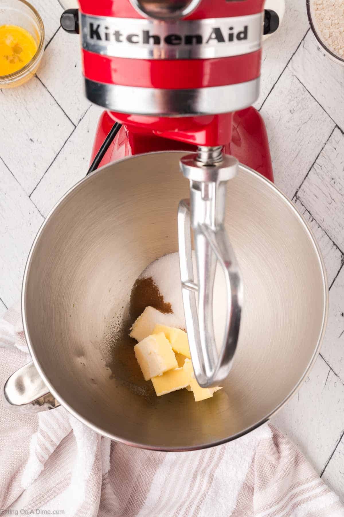 Top-down view of a red KitchenAid stand mixer with butter and sugar in the mixing bowl, ready to be mixed. A bowl with a yellow mixture and another bowl are visible in the background on a white surface.