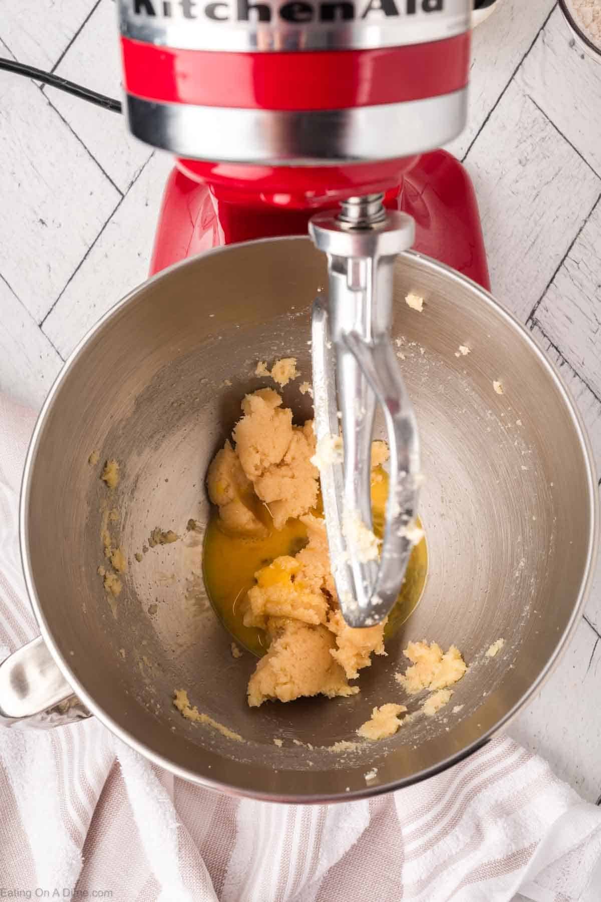 A red stand mixer with a paddle attachment is mixing sugar cookie dough and eggs in a stainless steel bowl on a white wooden surface.