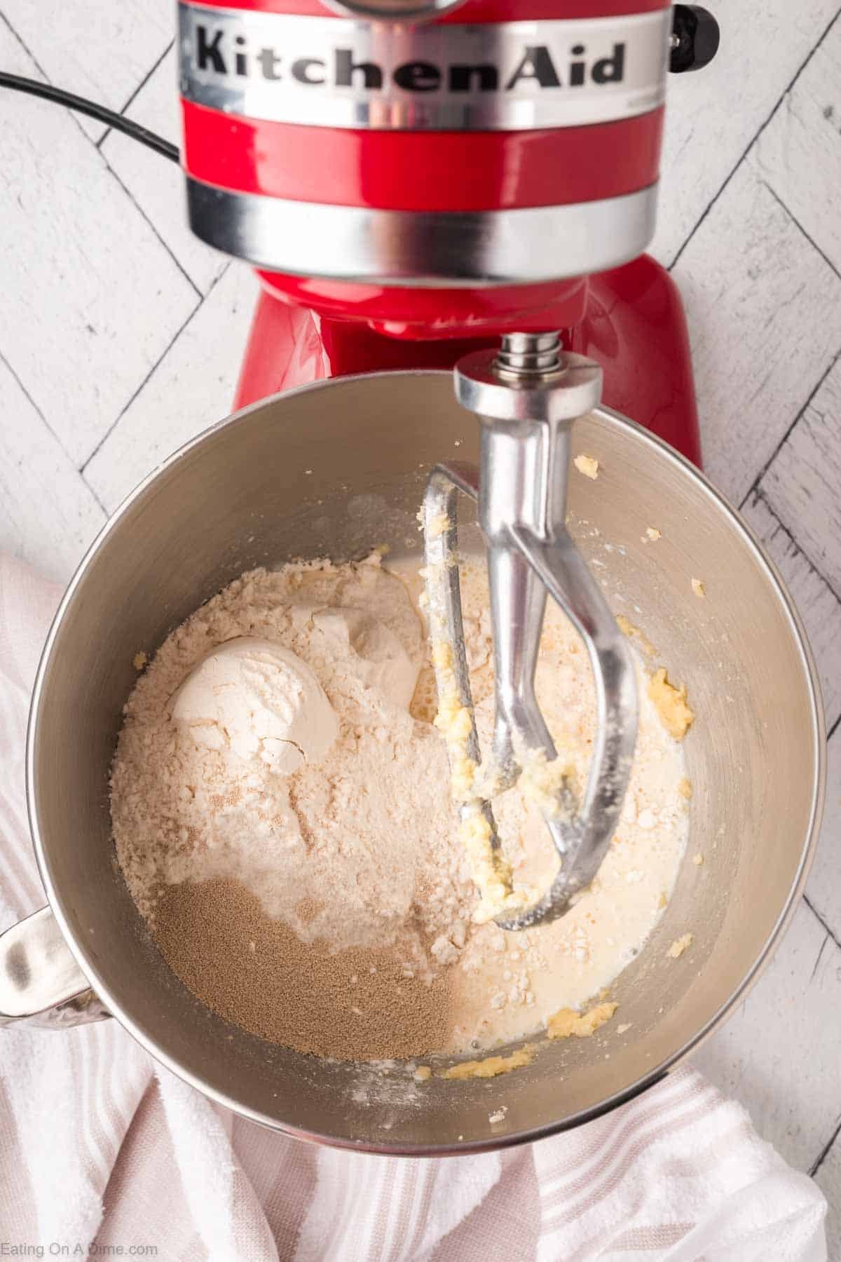 Top view of a red stand mixer with a metal bowl, containing flour, sugar, and yeast, partially mixed with a paddle attachment. A white kitchen towel is partially visible nearby.