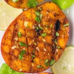 A close-up of a grilled mango half with a crisscross pattern, garnished with chopped cilantro and spices, surrounded by lime slices and fresh cilantro leaves highlights the vibrant appeal of grilled mango.