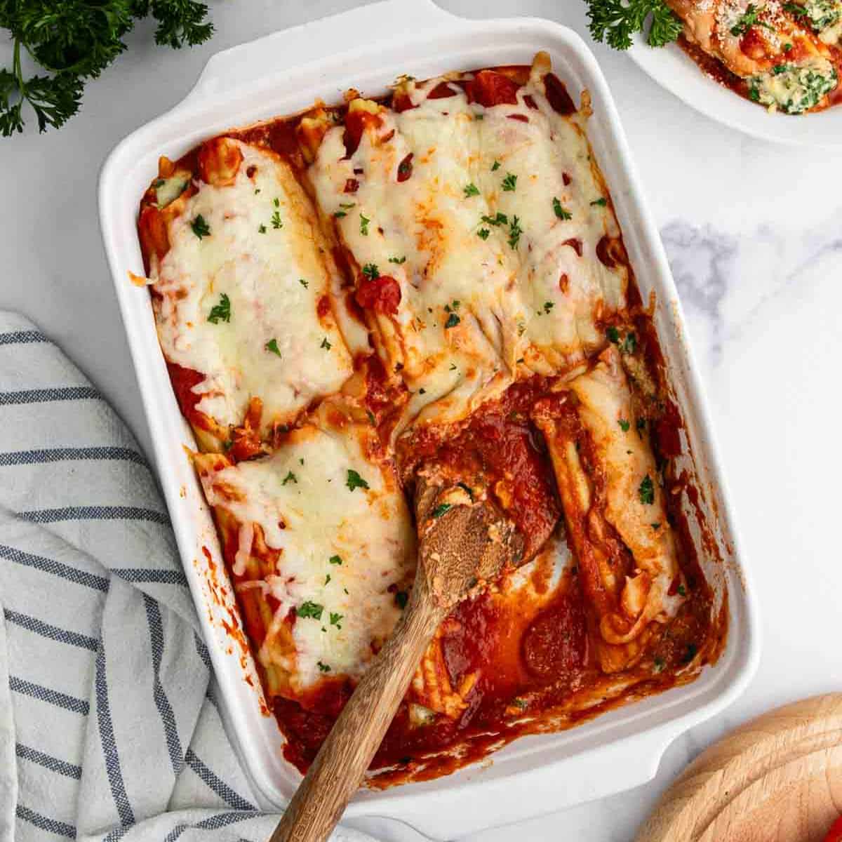 A baking dish with cheesy, baked lasagna rolls reminiscent of manicotti, topped with tomato sauce and sprinkled parsley; a wooden spoon rests inside, with one roll missing, on a marble surface next to a striped towel and parsley.