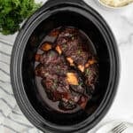 Slow Cooker Beef Ribs are slow-cooked to perfection, covered in a rich, dark sauce, and garnished with fresh herbs. Served in a black crockpot with parsley and mashed potatoes on the side for a comforting meal.