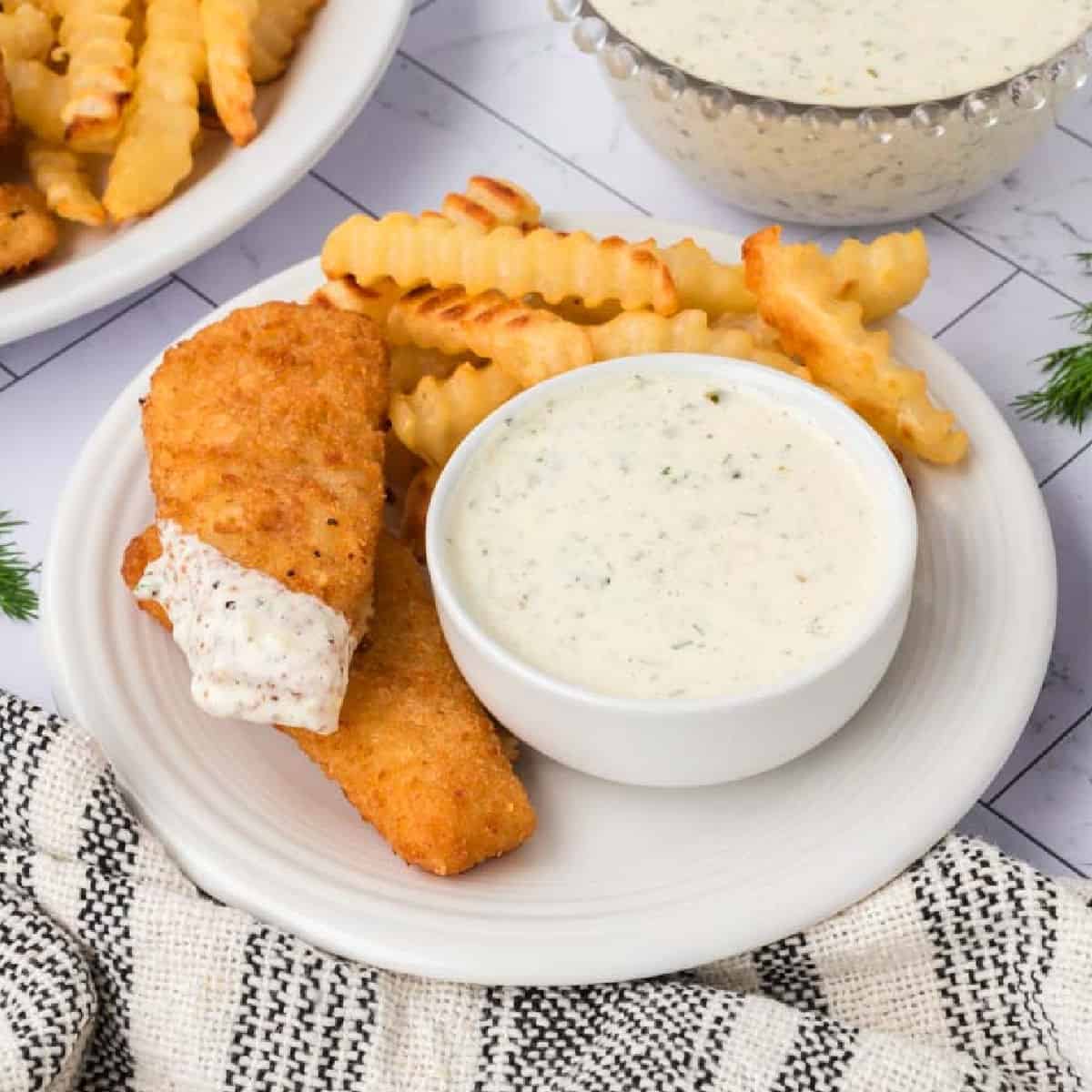 A plate with crinkle-cut fries, two breaded fish fillets—one topped with homemade tartar sauce from our Tartar Sauce Recipe—and a small bowl of creamy dipping sauce. A striped towel and more fries with dip are in the background.