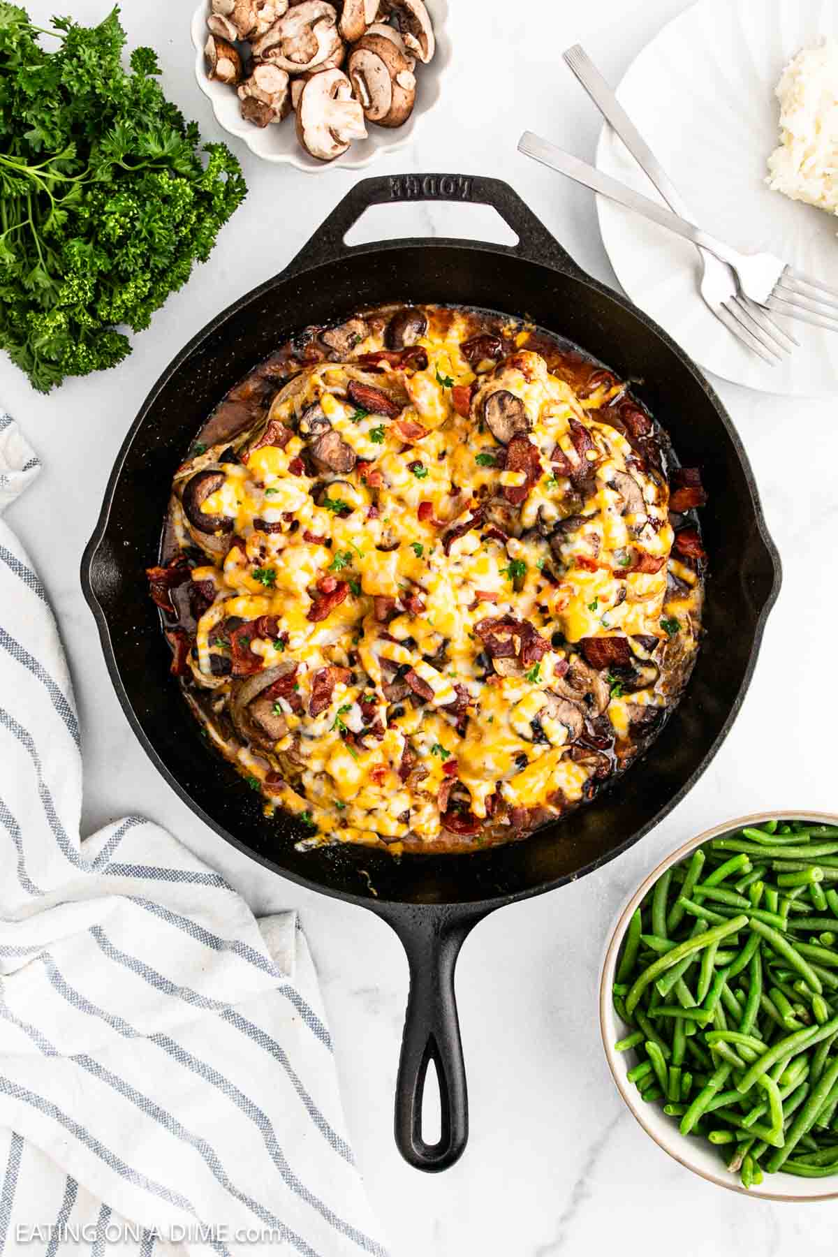 A skillet filled with Alice Springs Chickenโbaked chicken, mushrooms, melted cheese, and herbsโsits on a white countertop next to a bowl of green beans, fresh parsley, sliced mushrooms, and a striped towel.