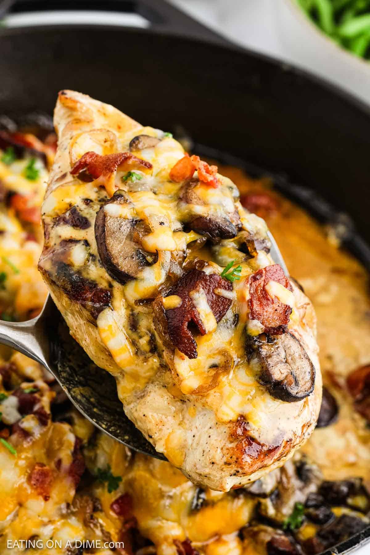 A close-up of Alice Springs Chicken: a cooked chicken breast topped with melted cheese, mushrooms, bacon bits, and herbs, being lifted from a skillet with a spoon.