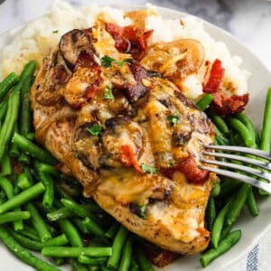 A plate of green beans and mashed potatoes topped with Alice Springs Chicken—baked chicken breast smothered in melted cheese, mushrooms, bacon pieces, and herbs—with a fork resting on the chicken.