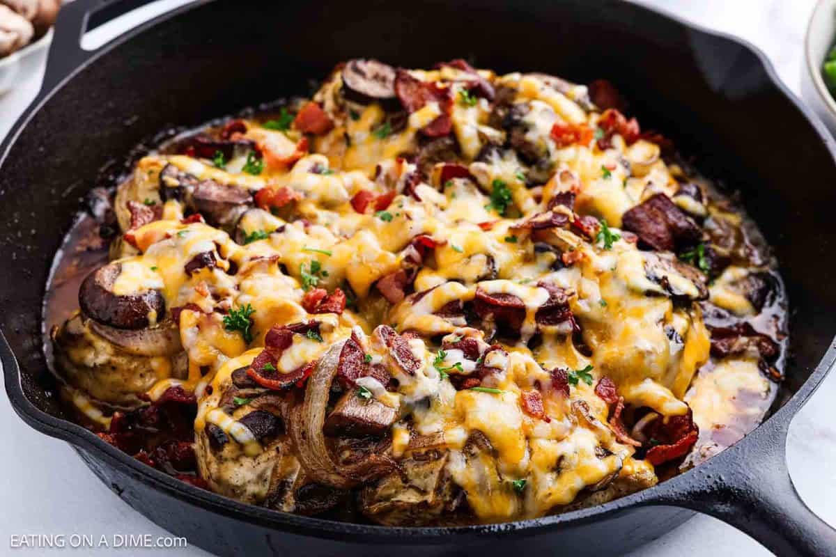 A cast iron skillet filled with Alice Springs Chickenโbaked chicken breasts topped with melted cheese, bacon pieces, mushrooms, and chopped parsley.