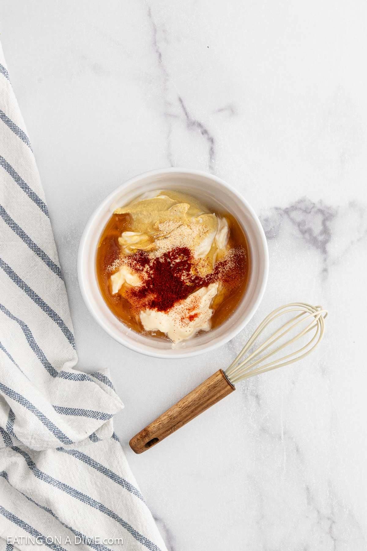 A white bowl containing mayonnaise, mustard, honey, garlic powder, and paprikaโthe perfect sauce for Alice Springs Chickenโsits on a marble surface next to a striped towel and a small whisk with a wooden handle.