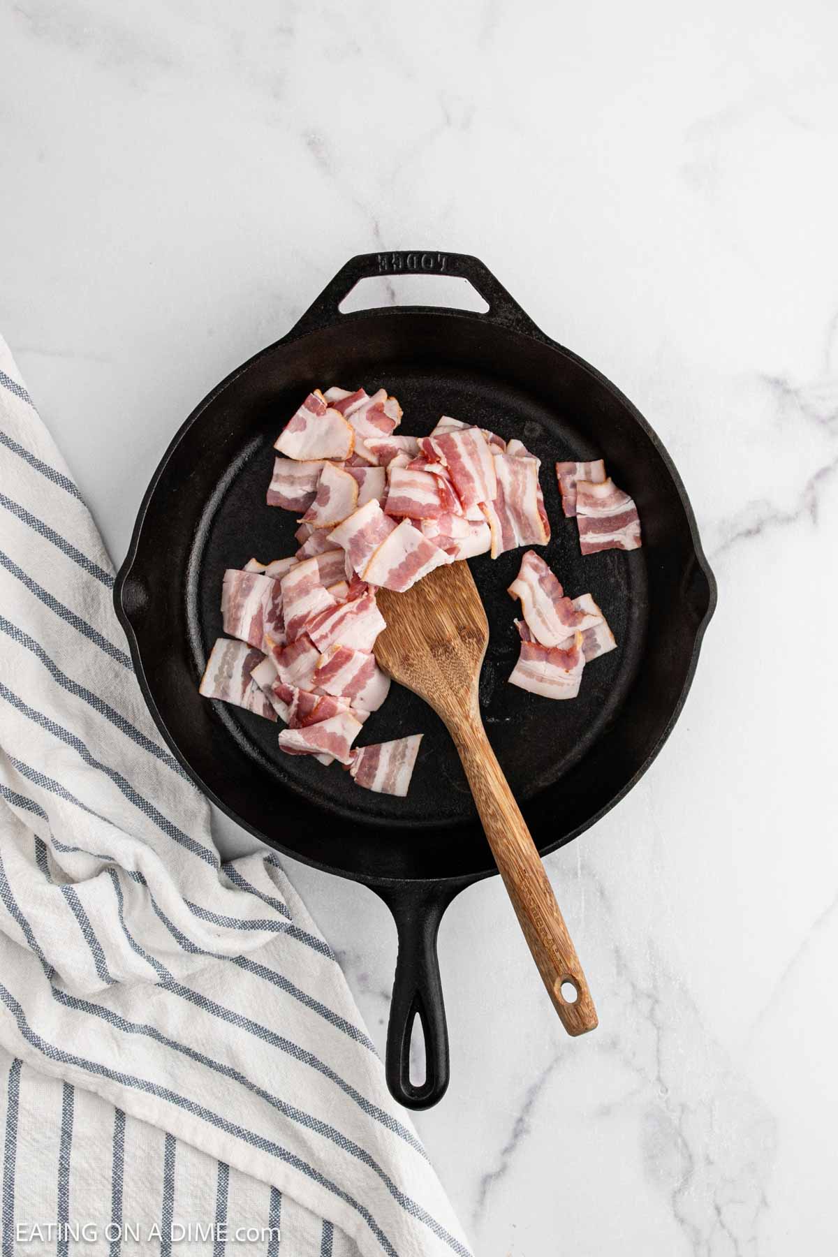 Cast iron skillet with raw, chopped bacon pieces and a wooden spatula on a white marble surfaceโperfect for starting Alice Springs Chickenโnext to a white and blue striped kitchen towel.