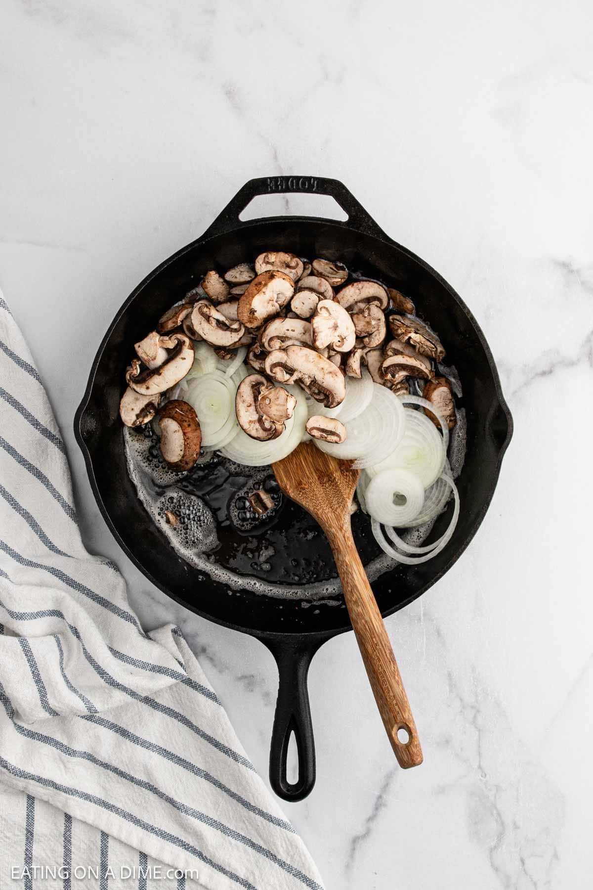 A cast iron skillet with sliced mushrooms and onions being sautรฉed, stirred with a wooden spoon, on a white marble surface next to a striped kitchen towelโperfect toppings for homemade Alice Springs Chicken.