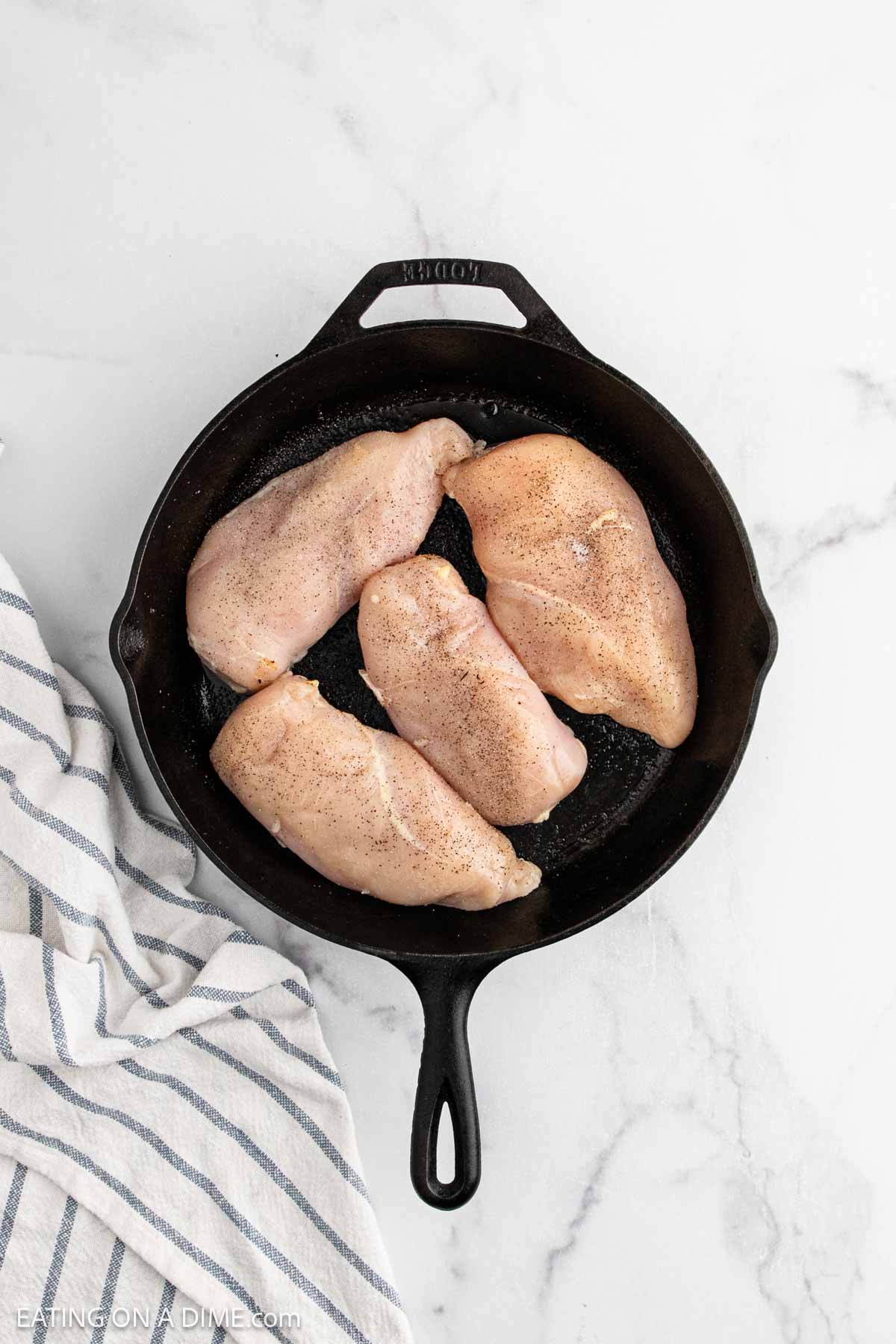 Four raw, seasoned chicken breasts arranged in a black cast iron skillet on a white marble surface, inspired by the flavors of Alice Springs Chicken, with a striped kitchen towel nearby.