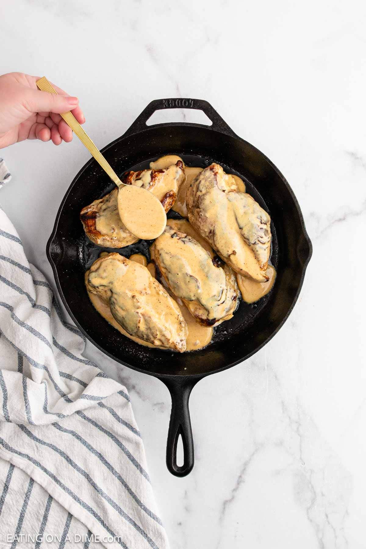 A hand spoons creamy sauce over four cooked Alice Springs Chicken breasts in a black cast iron skillet on a white marble surface, with a striped kitchen towel beside the skillet.