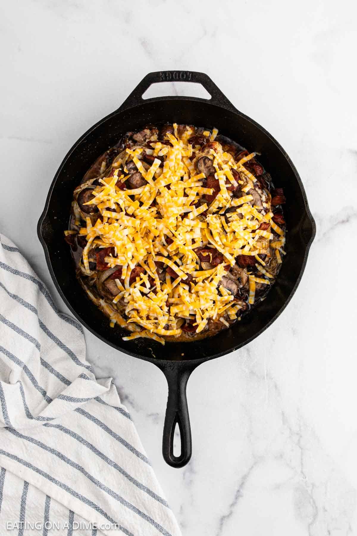 A black cast iron skillet filled with sliced cooked steak and vegetables, topped with a layer of shredded cheese, sits on a white marble surface next to a striped kitchen towel, reminiscent of the classic Alice Springs Chicken flavors.
