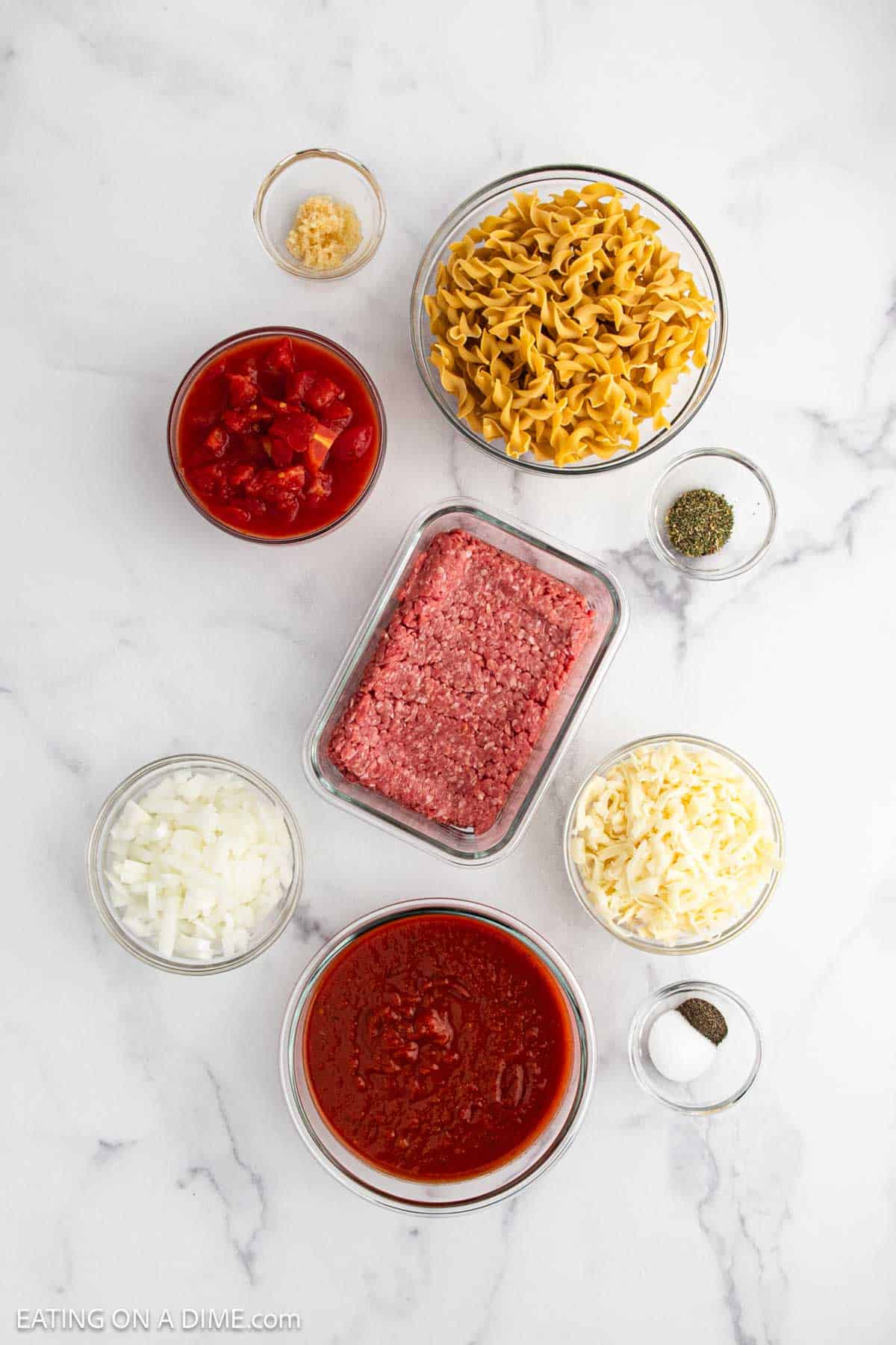 Top-down view of separate glass bowls containing uncooked egg noodles, ground beef, diced tomatoes, tomato sauce, chopped onion, shredded cheese, minced garlic, and small bowls of seasoning—perfect ingredients for a hearty Beef Casserole.