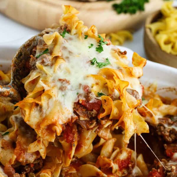 A close-up of a Beef and Noodle Casserole being served, featuring melted cheese, ground beef, tomato sauce, and egg noodles, garnished with chopped parsley.