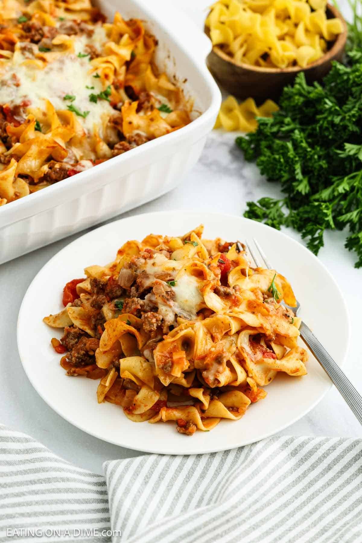 A plate of cheesy beef and noodle casserole with parsley sits on a white table next to a casserole dish, fresh parsley, uncooked noodles, and a fork.
