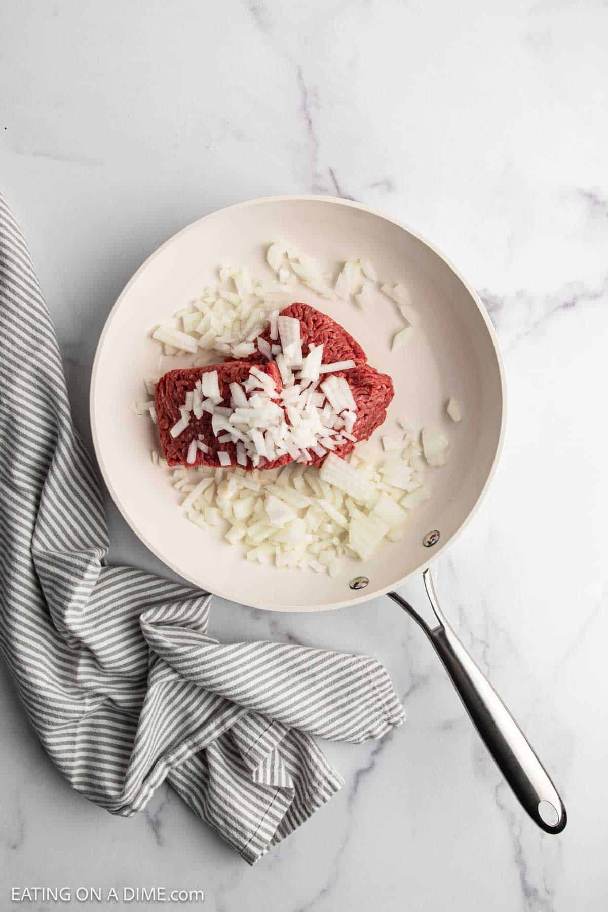 A white skillet on a marble surface holds raw ground beef and chopped onions—perfect beginnings for a hearty Beef Noodle Casserole. A gray and white striped kitchen towel rests beside the skillet.