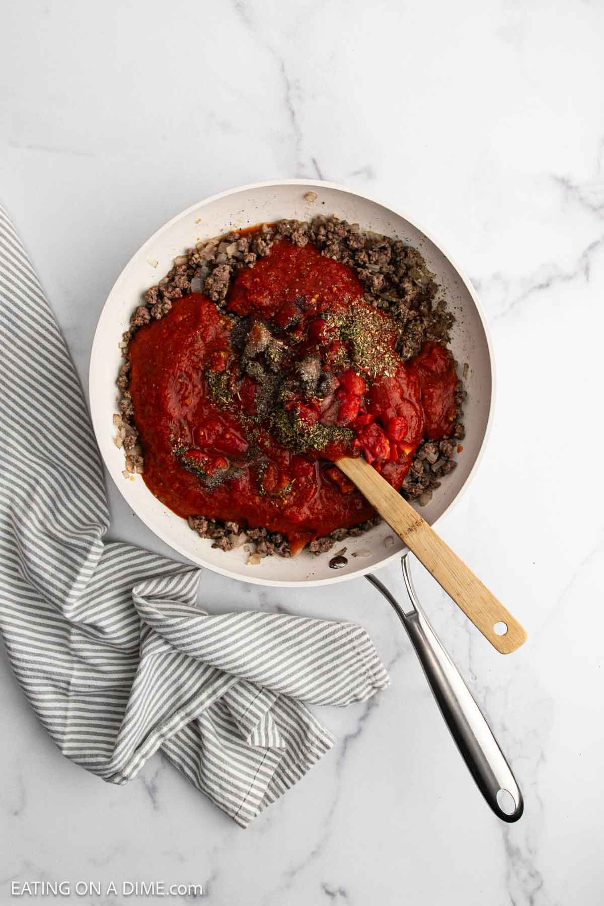 A skillet filled with cooked ground beef, tomato sauce, and herbs for a hearty beef casserole is being stirred with a wooden spoon, placed on a marble countertop next to a gray and white striped kitchen towel.