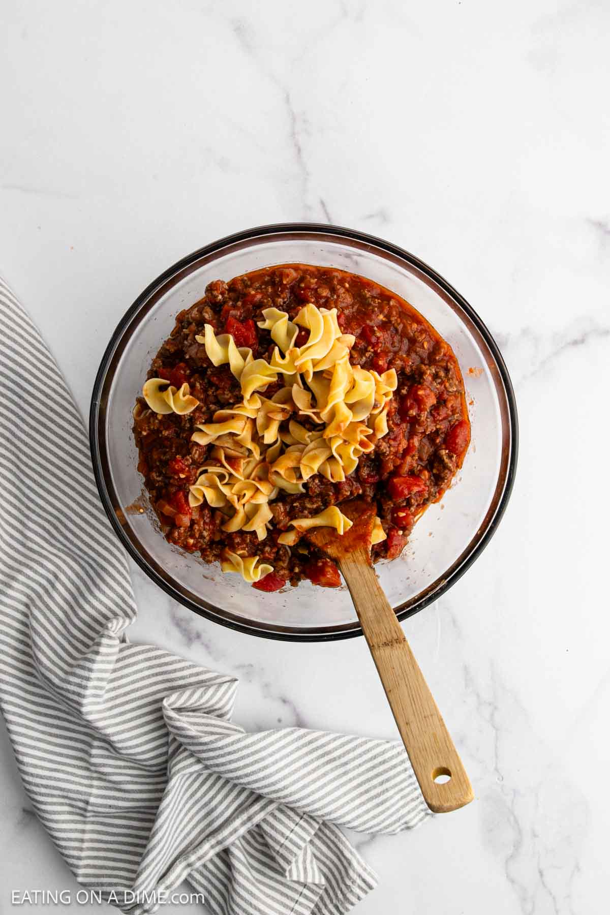 A glass bowl filled with a hearty beef and noodle casserole—cooked ground beef, tomato sauce, and uncooked egg noodles—rests on a white marble surface beside a gray striped cloth, with a wooden spoon inside.