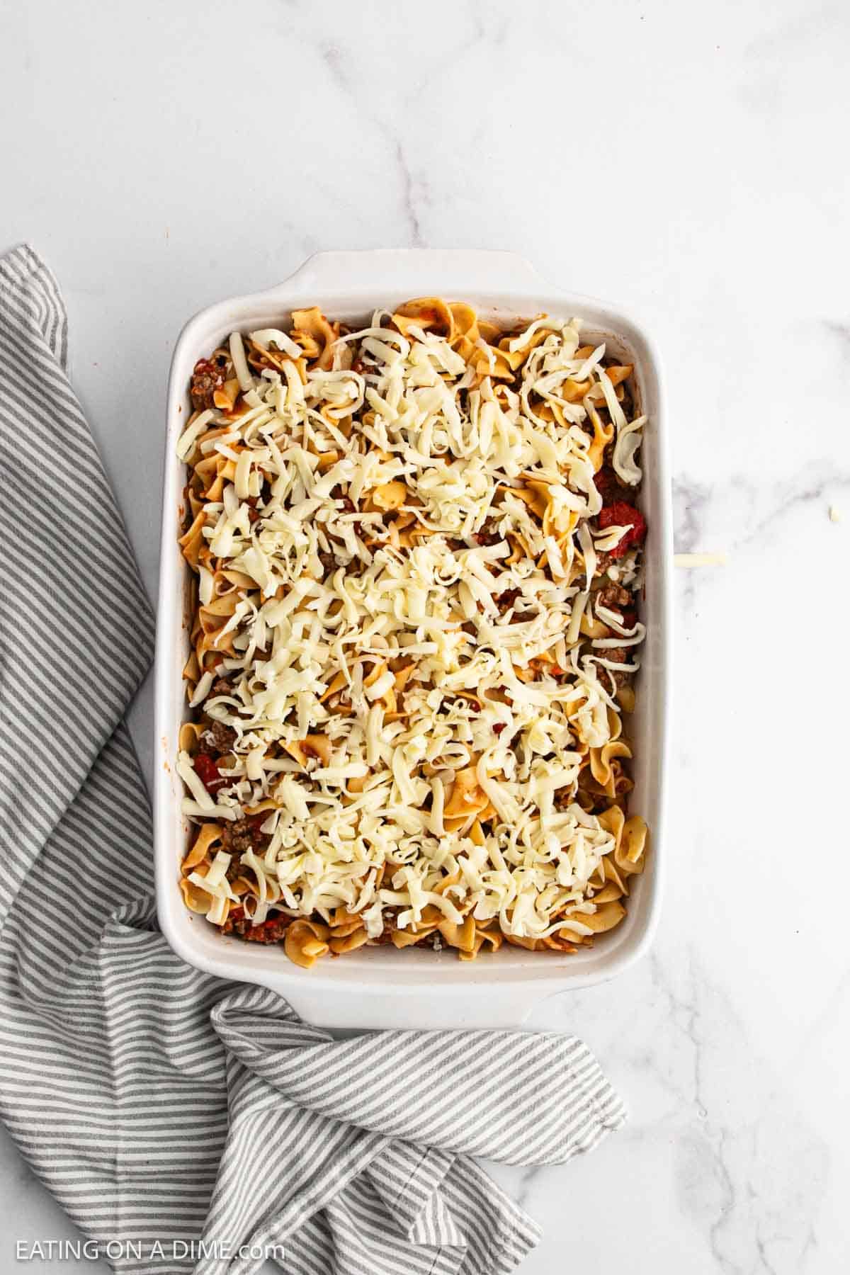 A rectangular baking dish filled with beef and noodle casserole, layered with ground meat, tomato sauce, and topped with shredded cheese sits on a white marble surface next to a gray and white striped cloth.