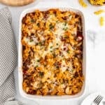 A baked Noodle Casserole filled with cheesy, saucy egg noodles and sprinkled with fresh herbs, placed on a white surface next to a striped towel and some uncooked noodles.