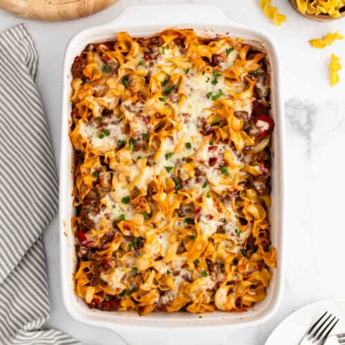 A baked Noodle Casserole filled with cheesy, saucy egg noodles and sprinkled with fresh herbs, placed on a white surface next to a striped towel and some uncooked noodles.