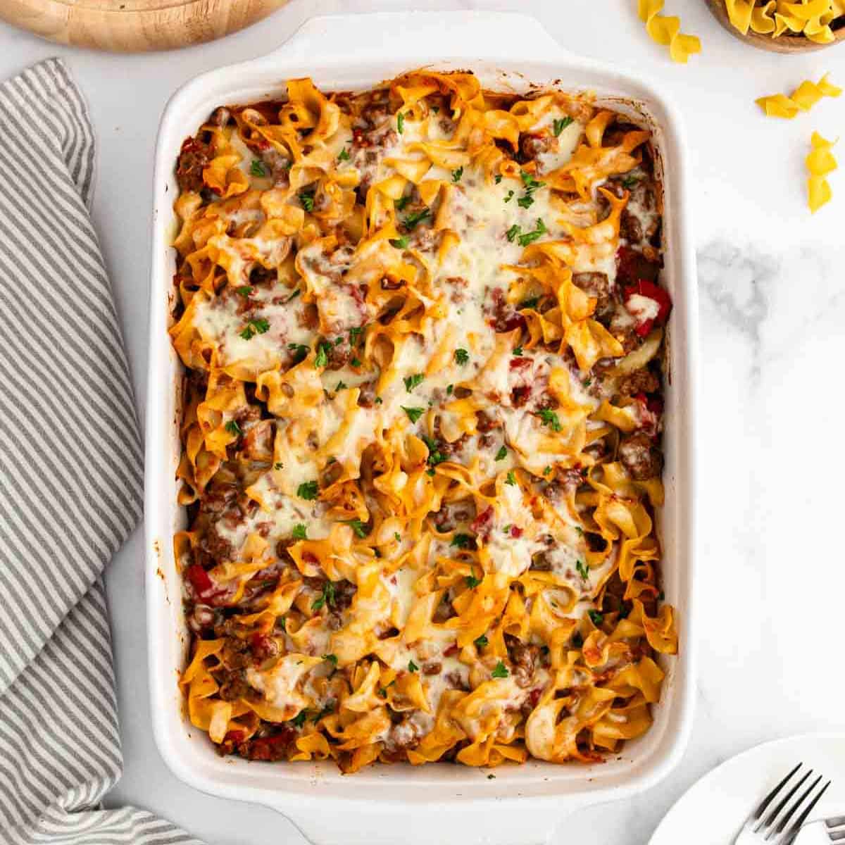 A baked Noodle Casserole filled with cheesy, saucy egg noodles and sprinkled with fresh herbs, placed on a white surface next to a striped towel and some uncooked noodles.