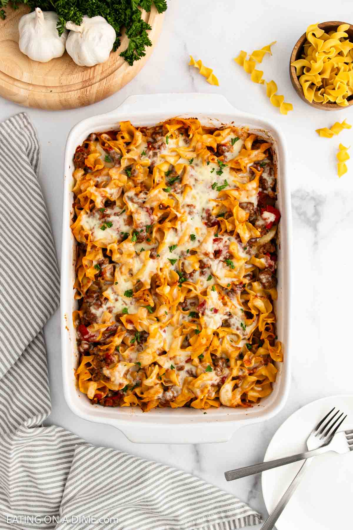 A white casserole dish filled with cheesy beef noodle casserole, topped with melted cheese and herbs, sits on a white marble surface beside a striped towel, utensils, uncooked noodles, parsley, and garlic.