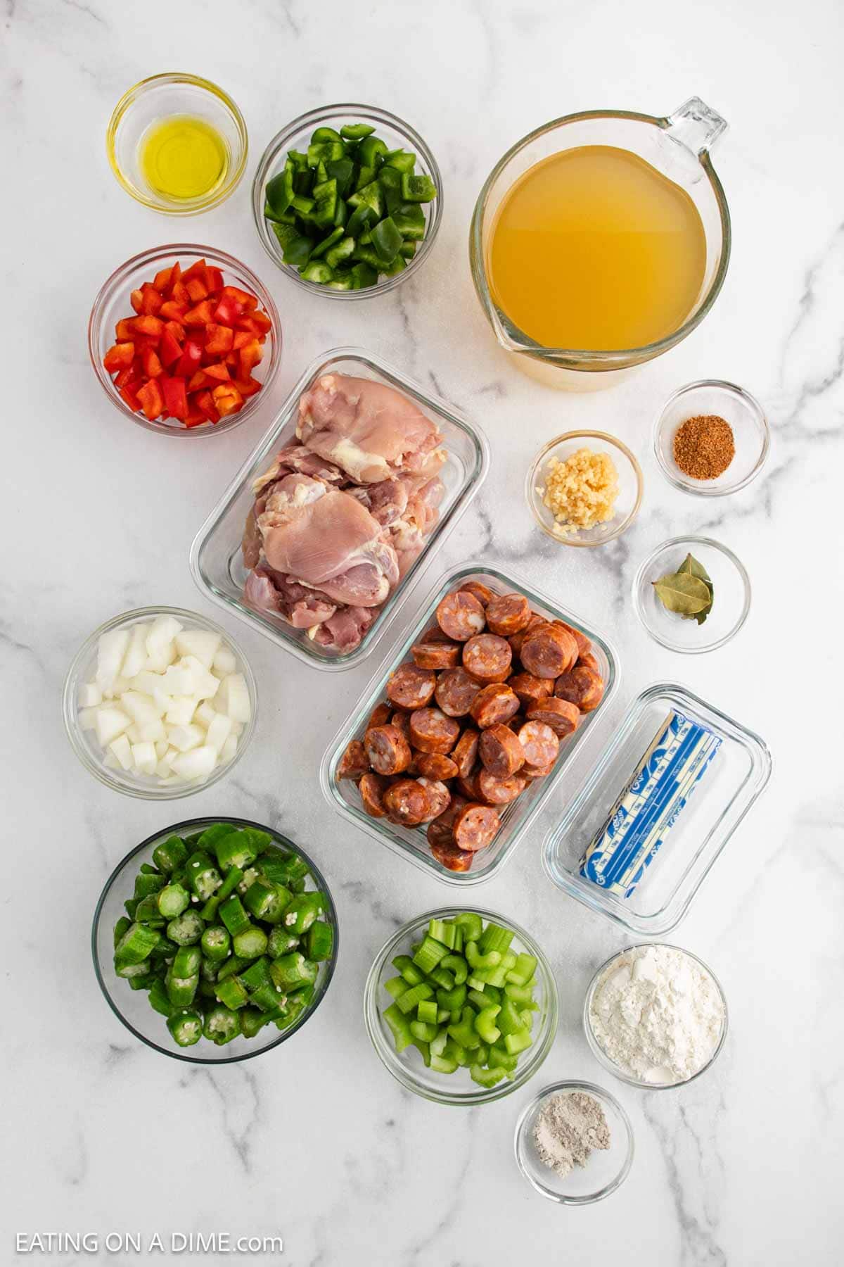 Top view of Chicken Gumbo Recipe ingredients on a white surface, featuring chopped onions, bell peppers, celery, okra, sausage, chicken thighs, minced garlic, seasoning, flour, butter, broth, and olive oil in small bowls and containers.