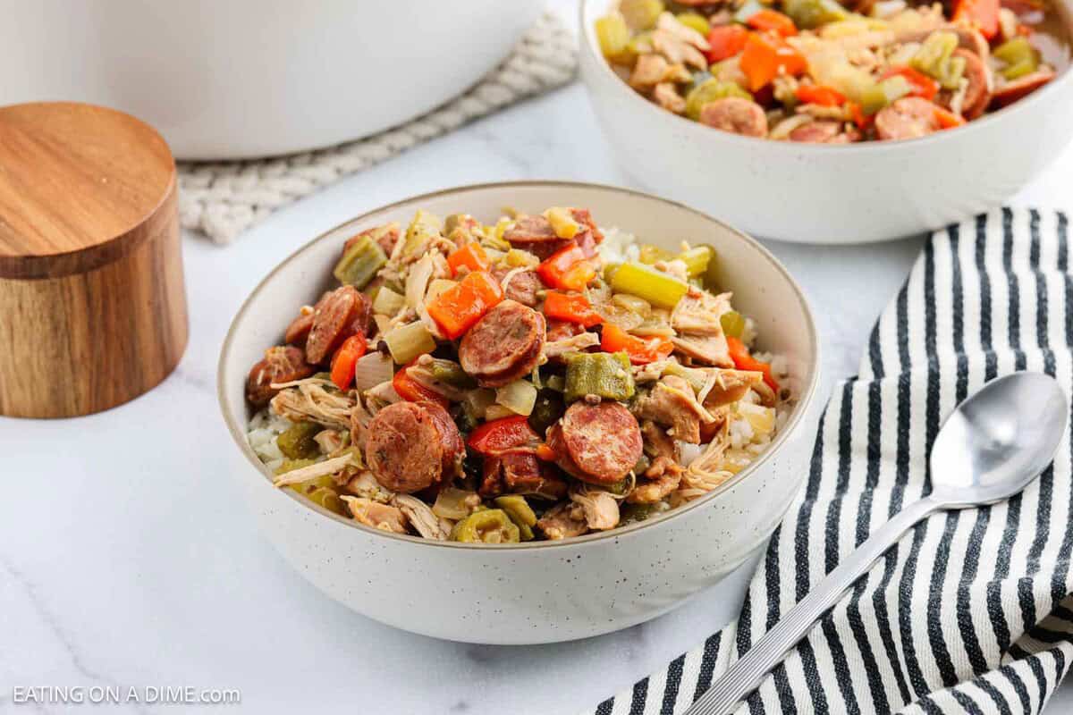 A bowl of rice topped with sliced sausage, shredded chicken, celery, bell peppers, and okra—a classic Chicken Gumbo Recipe. A silver spoon and a black-and-white striped napkin are beside the bowl, with another serving in the background.